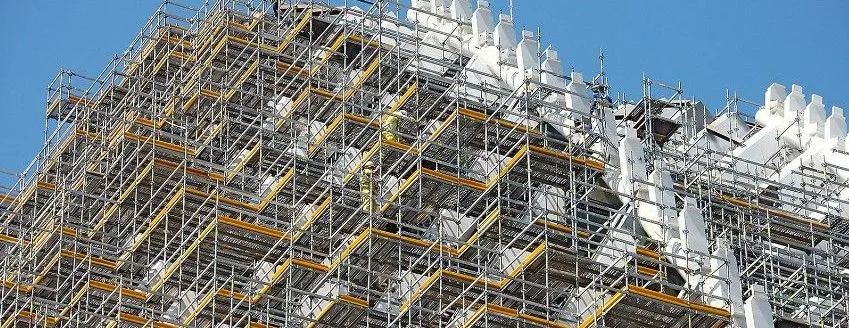 Primer plano de un edificio en construcción, donde se aprecian extensos andamios y trabajadores sobre la estructura, con un cielo azul despejado de fondo.